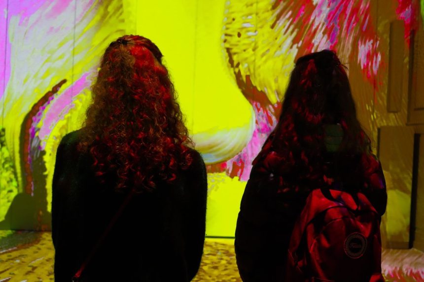 Duas mulheres observam uma obra de arte digital colorida em uma galeria de museu.