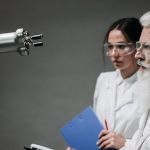 Dois pesquisadores usando jalecos e óculos de proteção analisam um braço robótico em um laboratório.