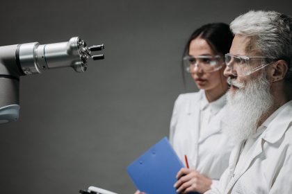 Dois pesquisadores usando jalecos e óculos de proteção analisam um braço robótico em um laboratório.