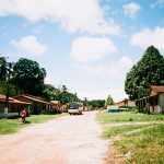 Ônibus trafega por uma estrada não pavimentada em uma comunidade rural, cercada por vegetação e moradias simples.