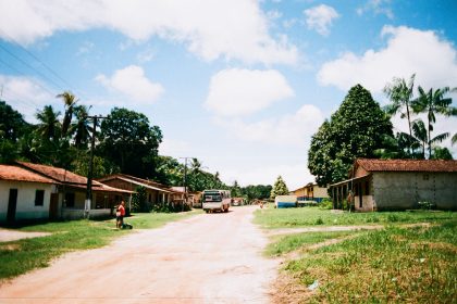 Ônibus trafega por uma estrada não pavimentada em uma comunidade rural, cercada por vegetação e moradias simples.