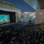 Público assiste a uma sessão de cinema ao ar livre durante a noite, sob um viaduto.
