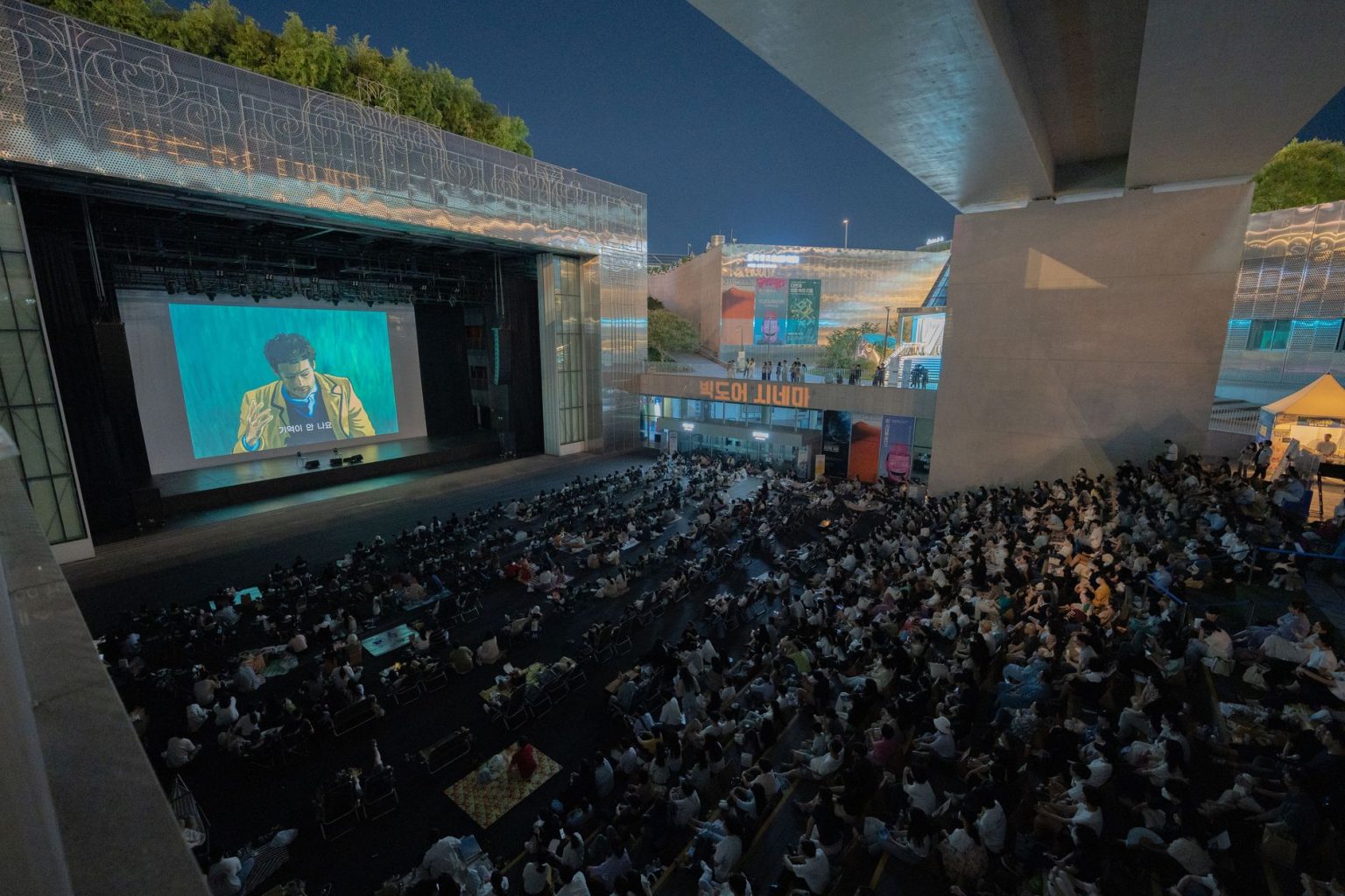 Público assiste a uma sessão de cinema ao ar livre durante a noite, sob um viaduto.